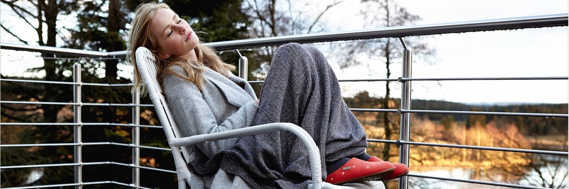 Woman in slippers curled up under blanket on wicker chair with scenic forest river in background